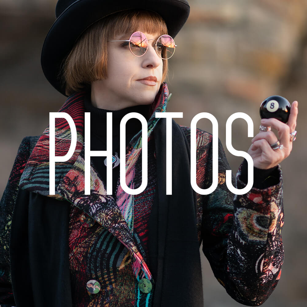 Photo of Jen Bateman wearing a multi-coloured tapestry coat and holding an 8-ball in Mill Ruins Park, Minneapolis, MN.